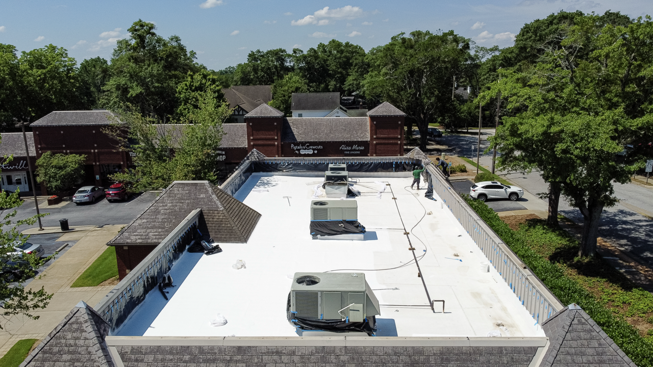 Construction workers installing a white roofing membrane on a commercial building with HVAC units, surrounded by trees and neighboring stores.