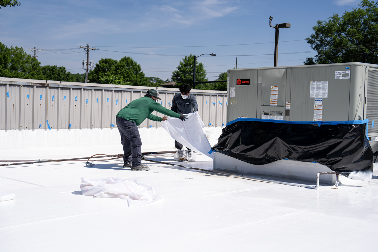 Two workers installing a white reflective coating on a building rooftop, one is helping the other with the material, with HVAC unit and safety equipment nearby, under a partly cloudy sky.
