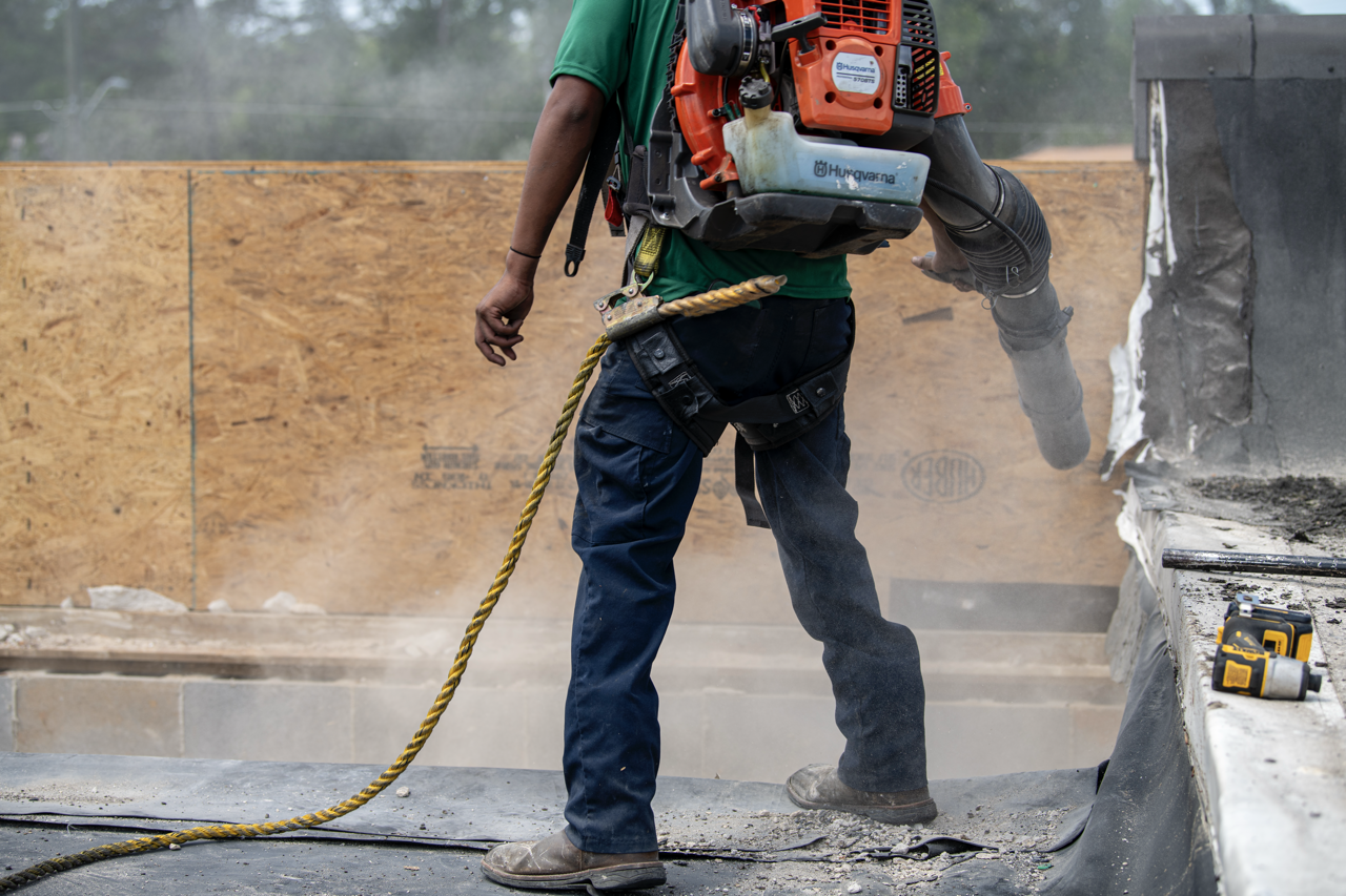 A construction worker operating a power tool, possibly a concrete grinder or demolition tool, on a building site with a partially damaged wall and construction equipment nearby.