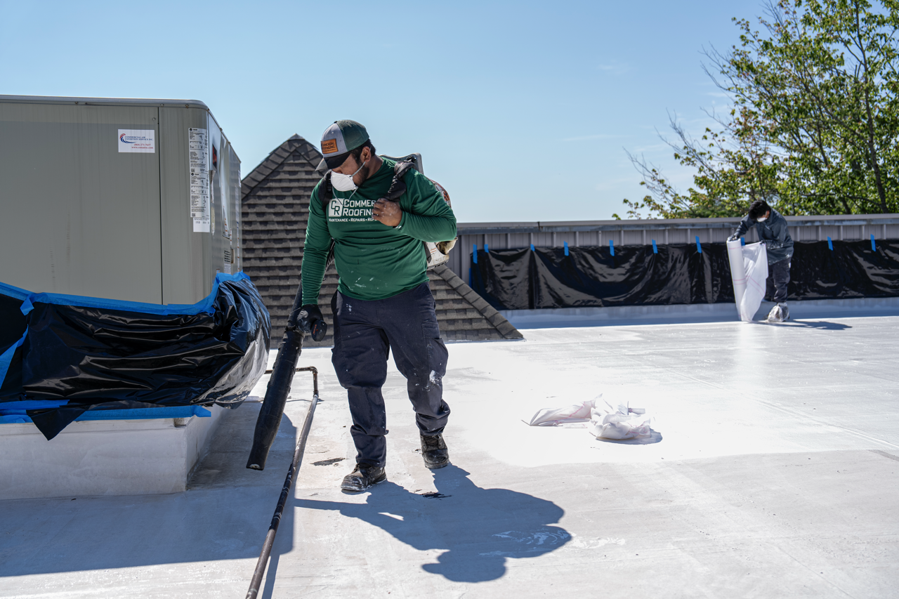 Two workers applying a white coating on a rooftop under a clear blue sky, with one worker in a green shirt and the other in dark clothing, both wearing masks and gloves.
