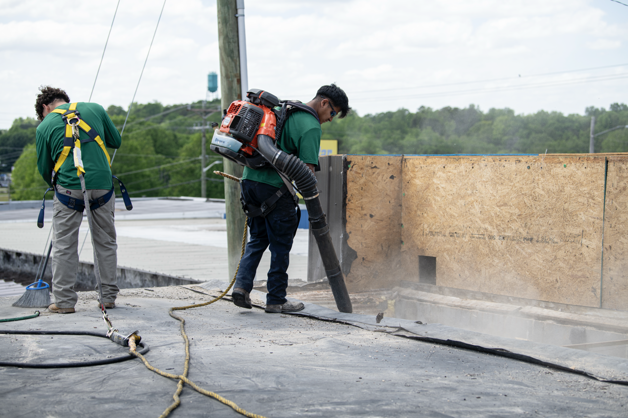 Two construction workers laying asphalt on a road. One is using a heavy-duty vacuum to remove debris, and the other is standing nearby with cleaning tools.