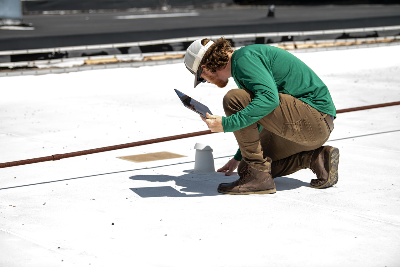 A man wearing a green long sleeve shirt, brown pants, and a white cap crouching on a rooftop, inspecting or working on a vent pipe with a tablet in his hand.