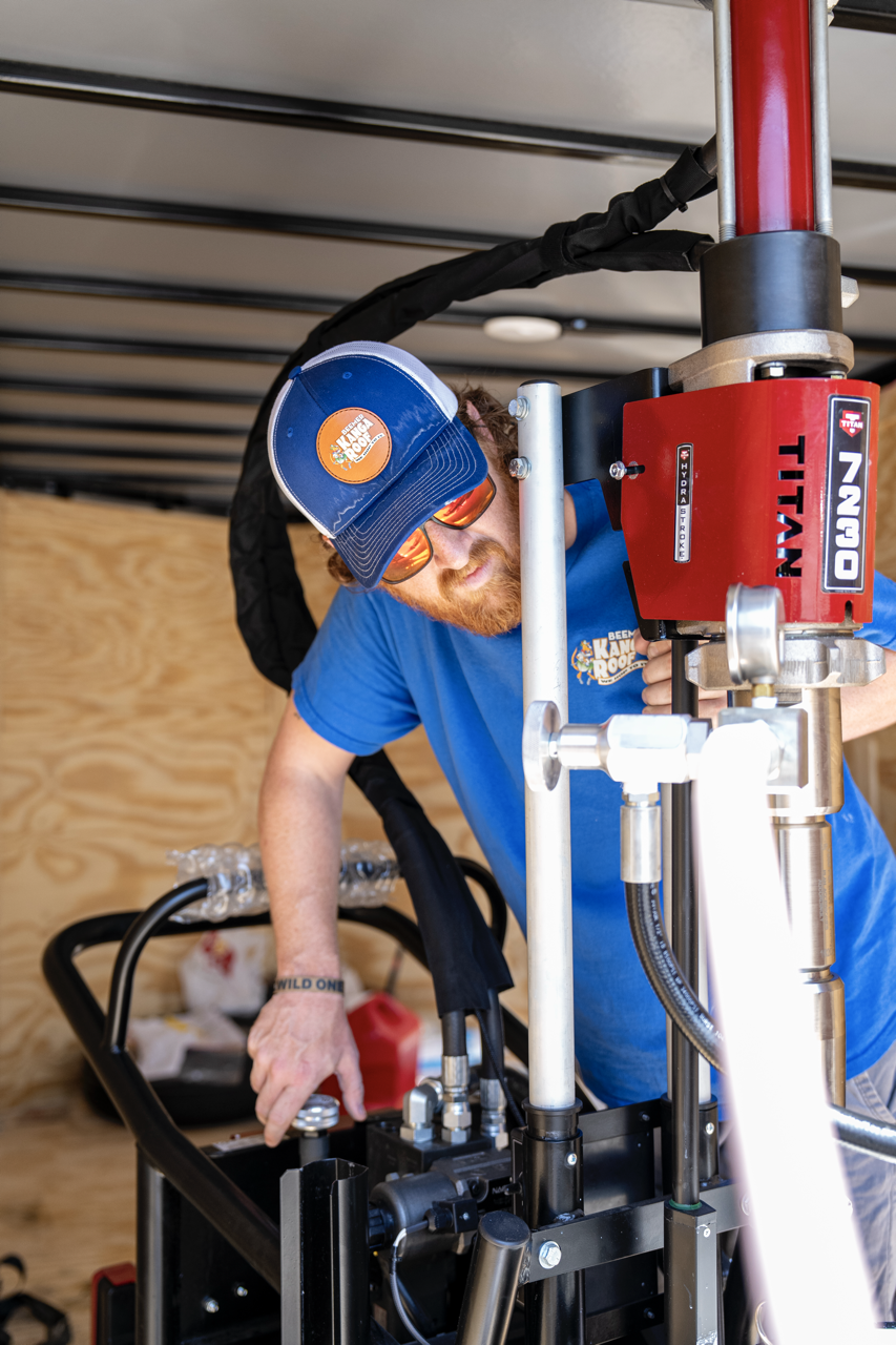 A man with a beard, wearing a blue T-shirt, a blue cap, and sunglasses works with hydraulic machinery inside a wooden structure.