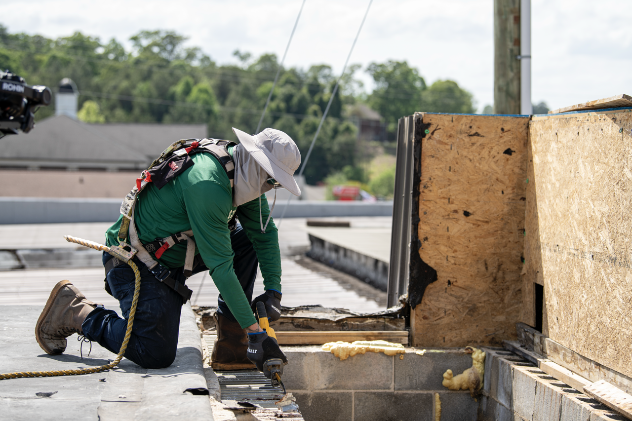 A construction worker wearing a hat, gloves, and safety harness kneels on a roof removing old roofing material with a power tool, with plywood sheathing nearby.