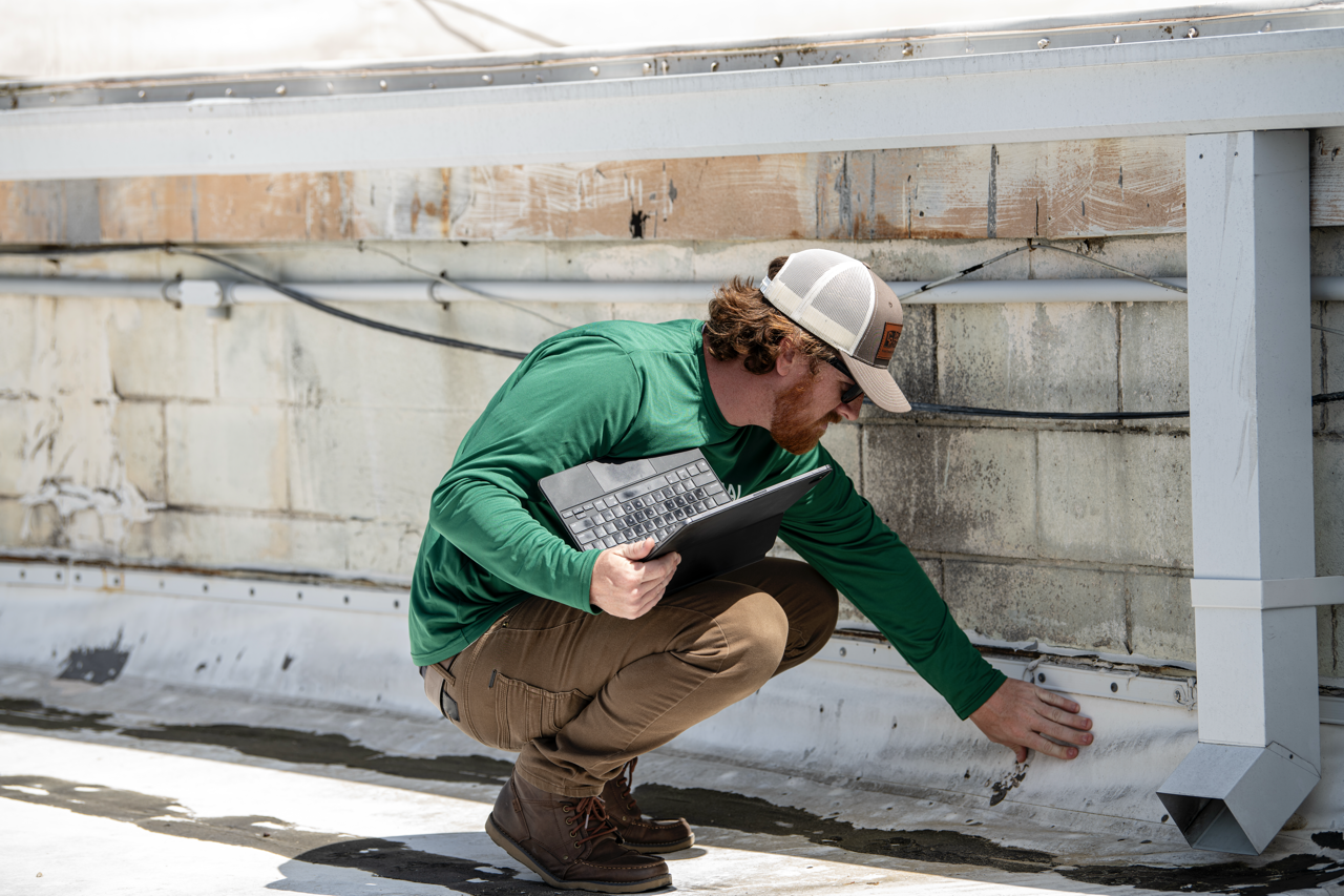 A man in a green shirt and baseball cap inspecting the base of a building's exterior wall with a laptop under his arm.