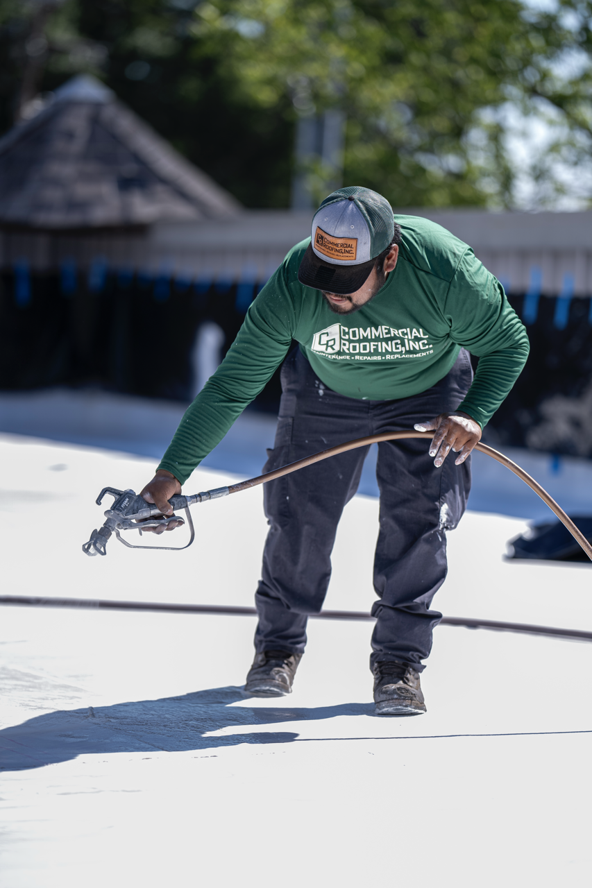 A worker in a green long-sleeve shirt and cap sprays a white waterproof coating on a flat roof.