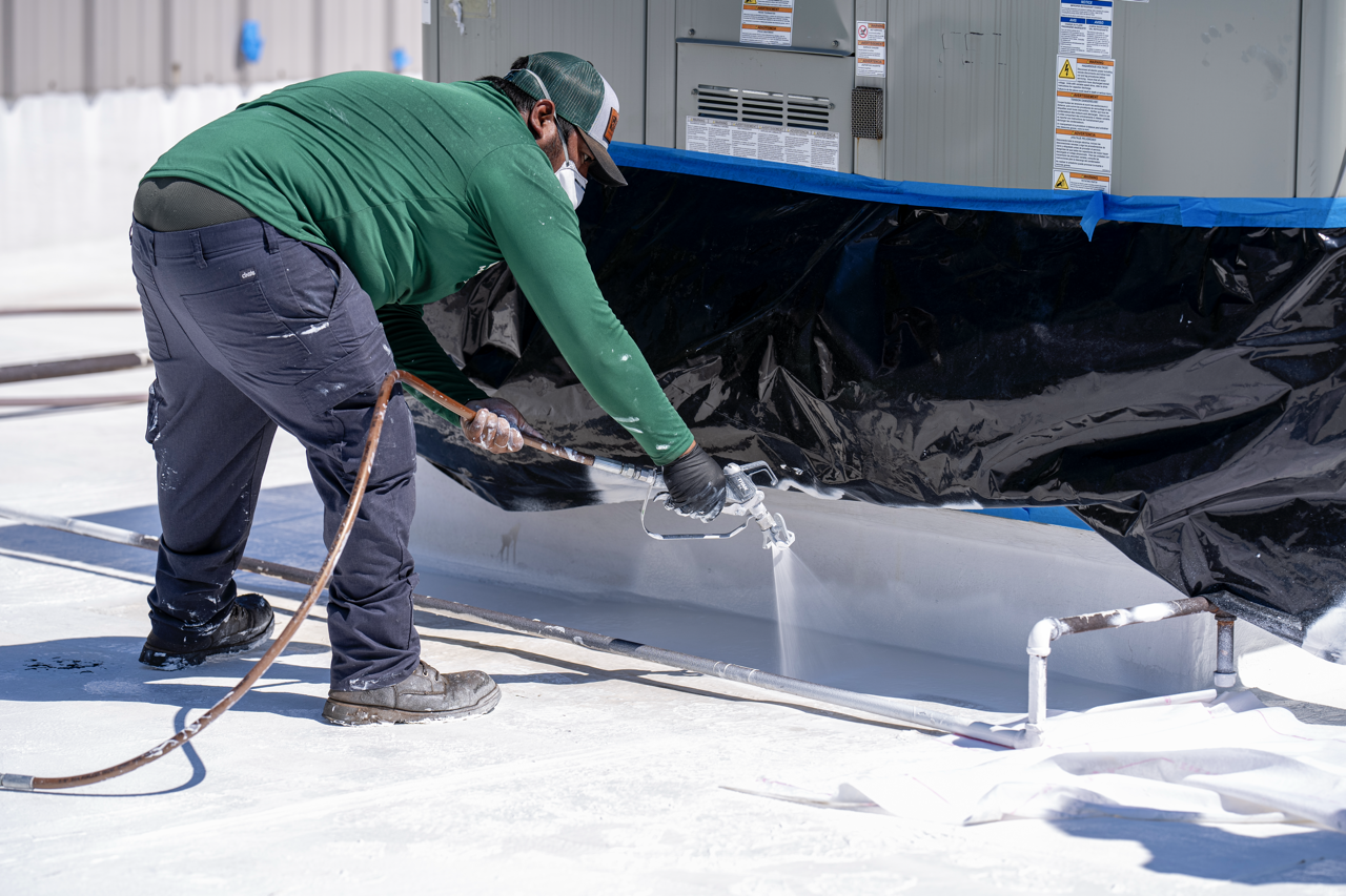 A worker is pressure washing the underside of a large outdoor air conditioning unit on a rooftop.