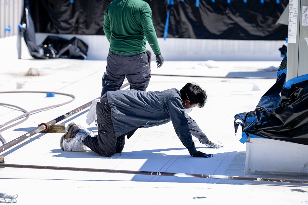 Two workers on a rooftop installing waterproofing membrane, one kneeling and the other standing, both wearing masks and gloves, with construction materials and equipment around them.