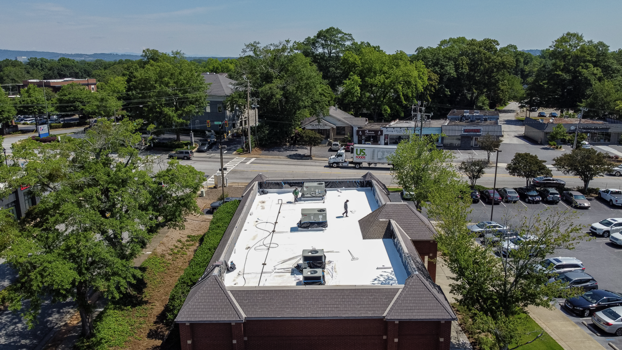 Aerial view of a commercial building with a flat white roof, two workers, and HVAC units, surrounded by trees, parking lot, and street with vehicles.