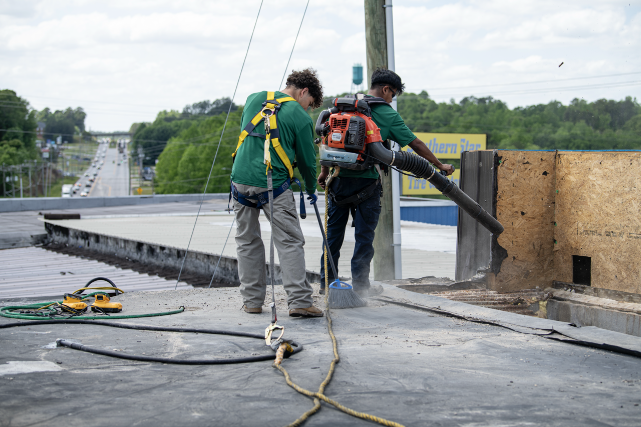 Two workers are on a rooftop, using a leaf blower to clear debris, with tools and safety harnesses visible.