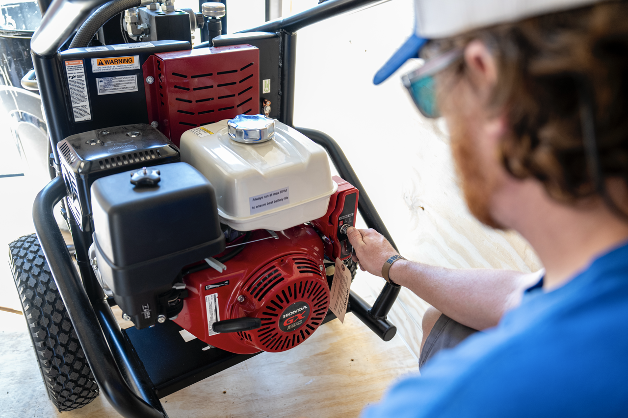 A person wearing a blue shirt, cap, and safety glasses operating a red Honda GX390 portable generator on a wooden surface indoors.