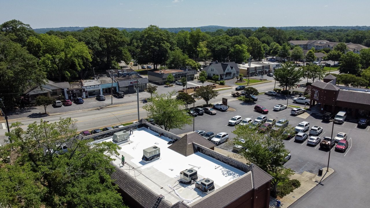 Aerial view of a small town with shops, parked cars, trees, and a building with a white roof under a clear sky.