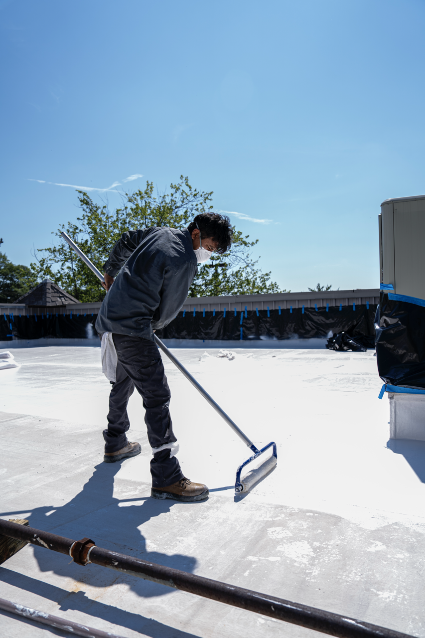 A person wearing a mask and work clothes is smoothing a white surface on a rooftop using a long-handled tool on a bright, sunny day.