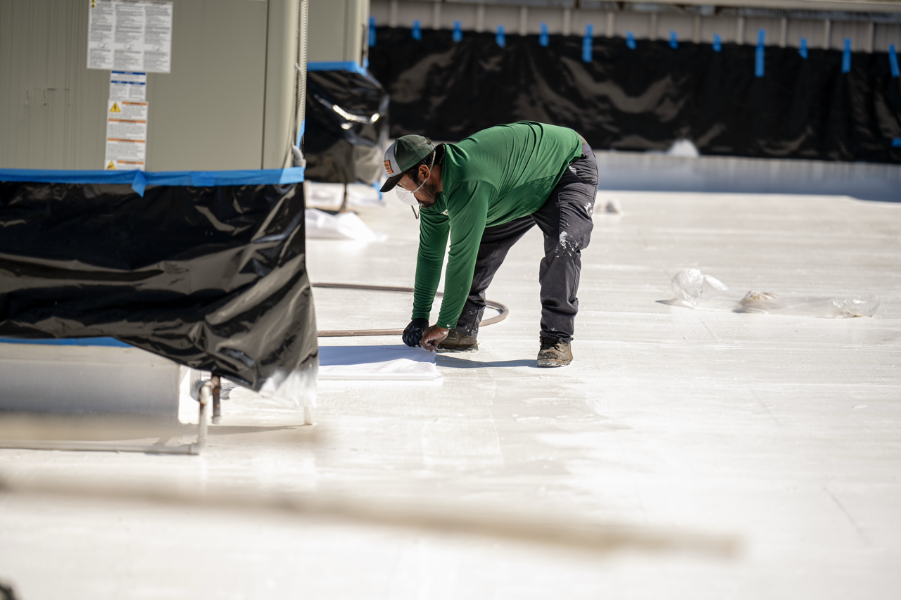 A person wearing a green long-sleeve shirt, gray pants, a hat, and a face mask is working on the roof of a building, handling tools near an HVAC unit.