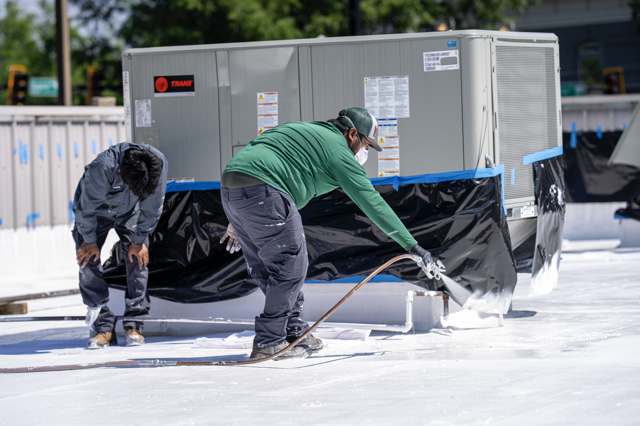 Two workers wearing masks and gloves spray foam insulation around an air conditioning unit on a rooftop.