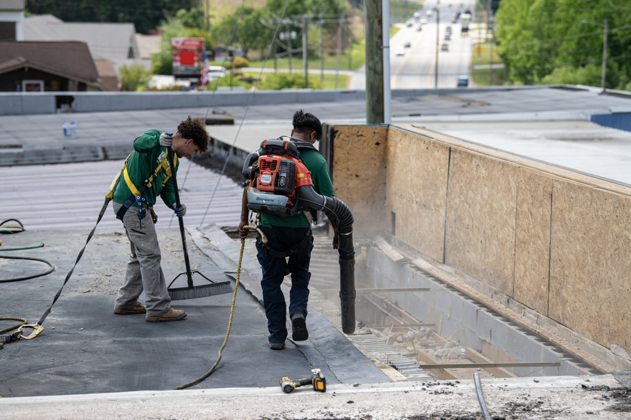 Two construction workers on a rooftop, one using a broom and the other operating a vacuum pump to remove debris from an open section of the roof, with a city street and trees in the background.