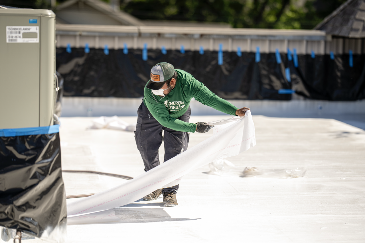 A construction worker wearing a green shirt, cap, and face mask installs a white waterproof membrane on a flat roof.