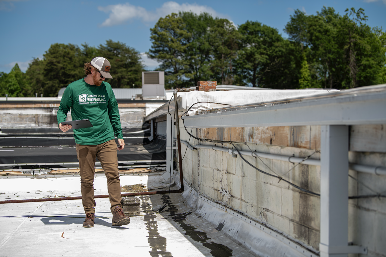 A man inspecting a commercial roof on a flat building, wearing a green long-sleeve shirt, beige pants, boots, and a trucker hat, holding a tablet computer, with trees and a blue sky in the background.