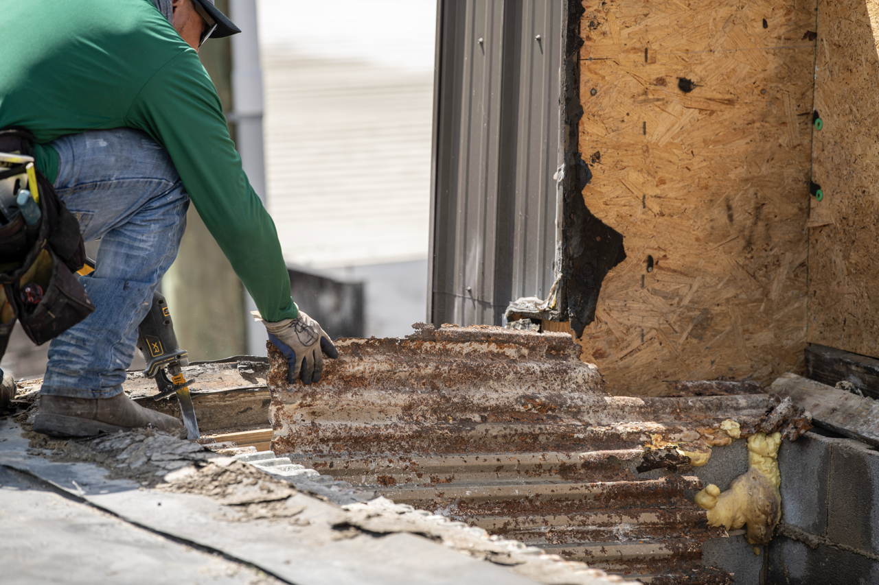 Construction worker removing rusted corrugated metal siding on a building with insulation and plywood wall behind.