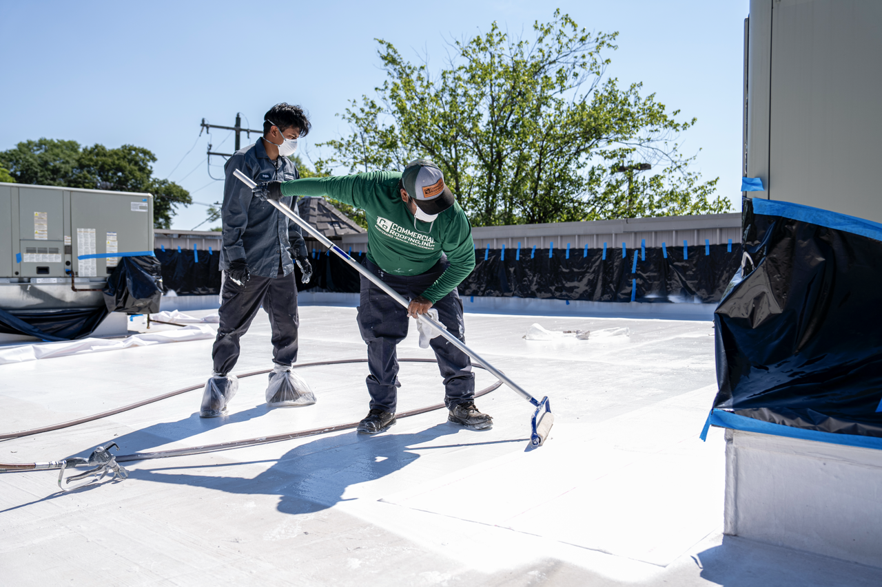 Two workers applying a white coating to a flat roof, with sunlight and a tree visible in the background.