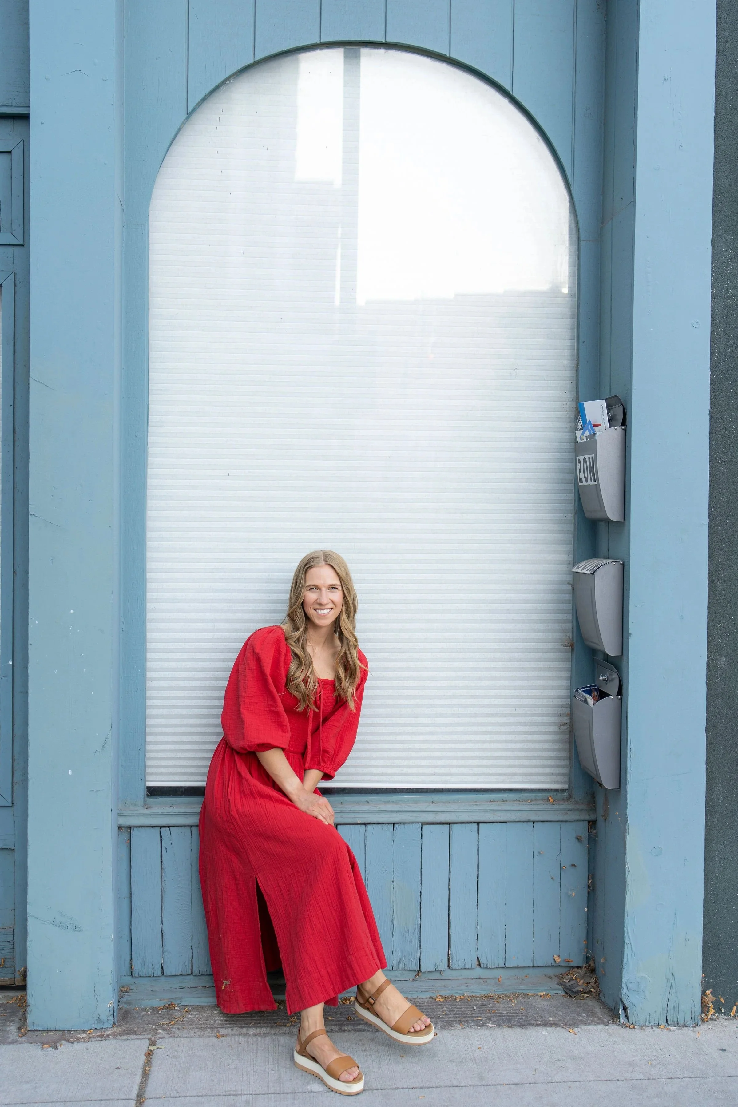 A woman with long wavy blonde hair, smiling, wearing a red dress and tan platform sandals, sitting on a blue window sill in front of a large white garage door with mailbox and mail on the right side.