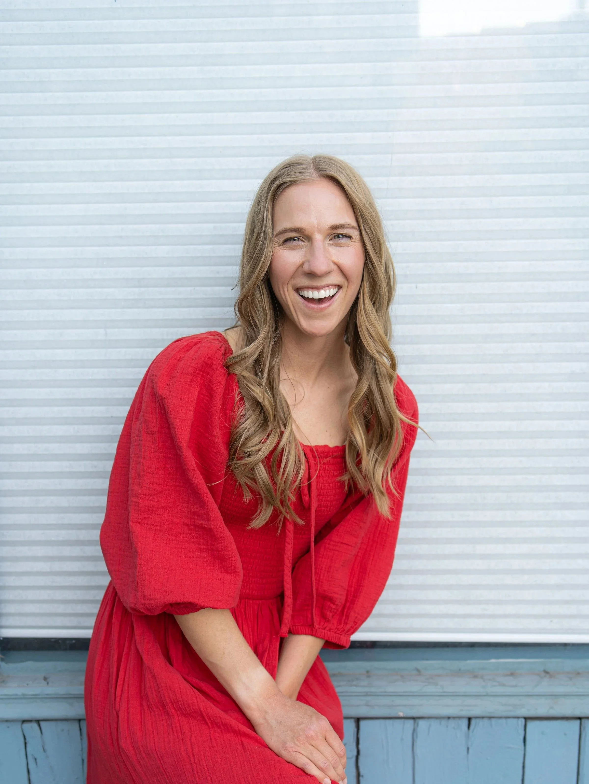 A woman wearing a red dress, smiling, with long blonde hair, sitting in front of a white, horizontally-striped background.