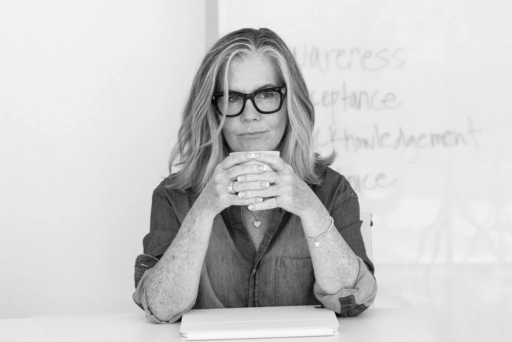 A woman with glasses and shoulder-length hair sits at a table, holding a cup with both hands. She is wearing a denim shirt and has a notebook in front of her. In the background, there is a whiteboard with handwritten notes that are partially visible.
