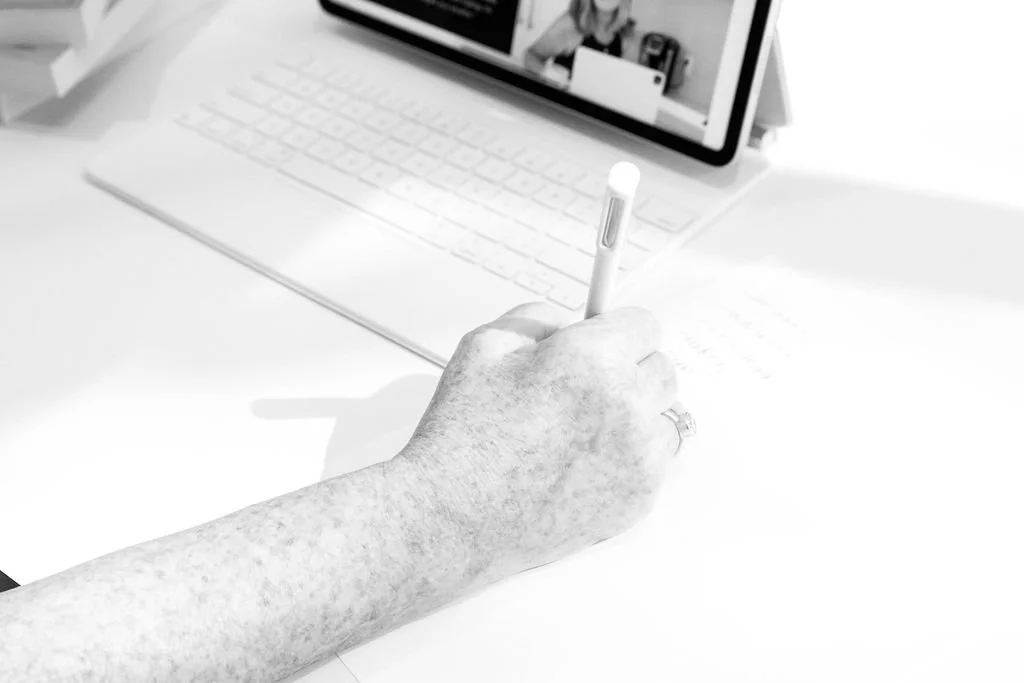 A person’s hand with a ring on the finger writing with a stylus on paper, with a tablet or laptop in the background on a white desk.