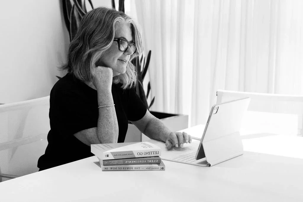 A woman with glasses and shoulder-length hair sitting at a desk, looking at a tablet, with three books stacked in front of her, in a bright room with curtains and a plant in the background.