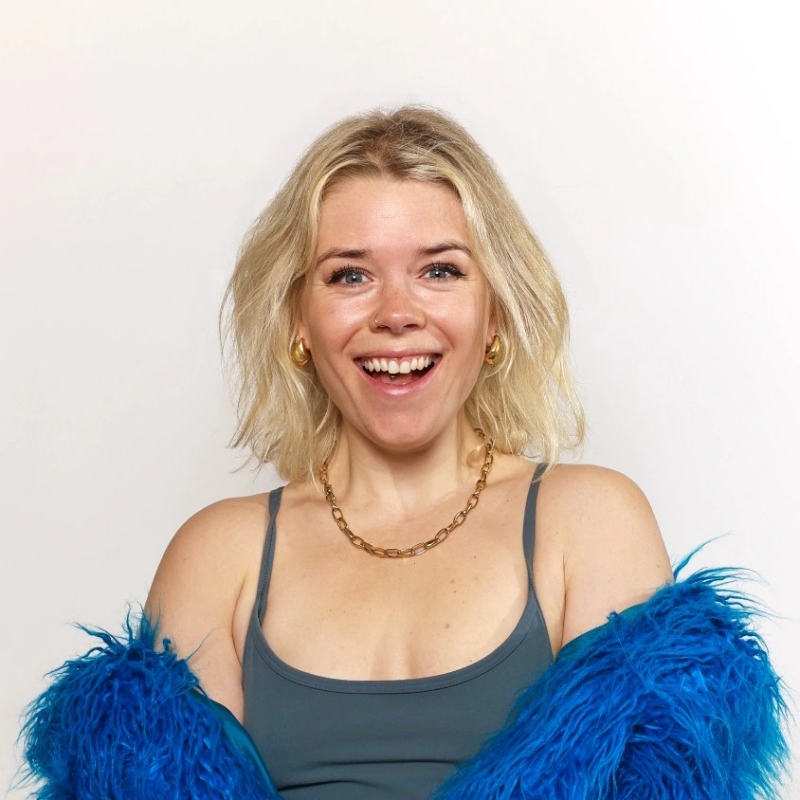 A smiling woman with blonde, wavy hair wearing a gray tank top, gold jewelry, and a blue feathered jacket against a plain white background.