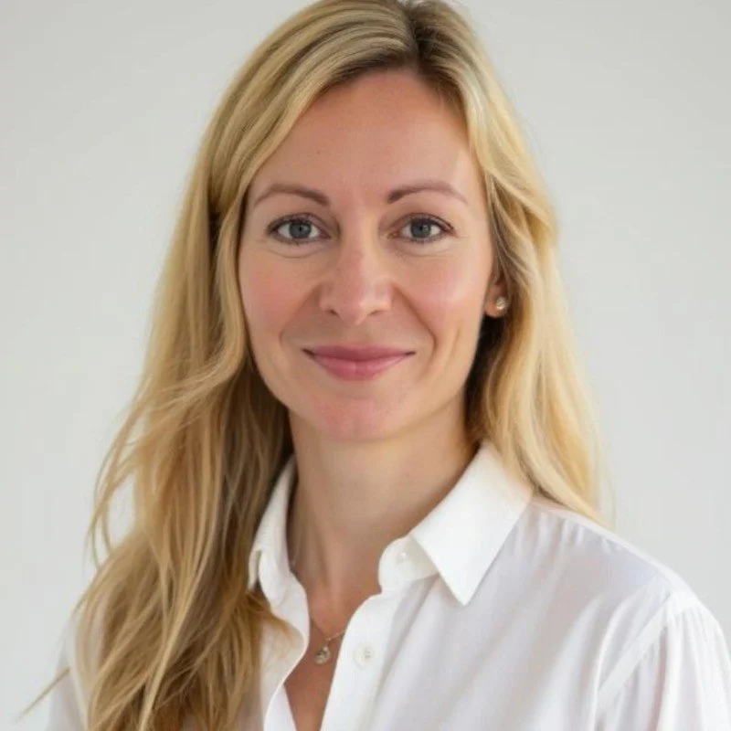 Portrait of a woman with blonde hair, blue eyes, wearing a white collared shirt and earrings, smiling against a plain background.