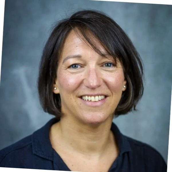 A woman with short dark brown hair, wearing a dark blue collared shirt, smiling, with a gray background.
