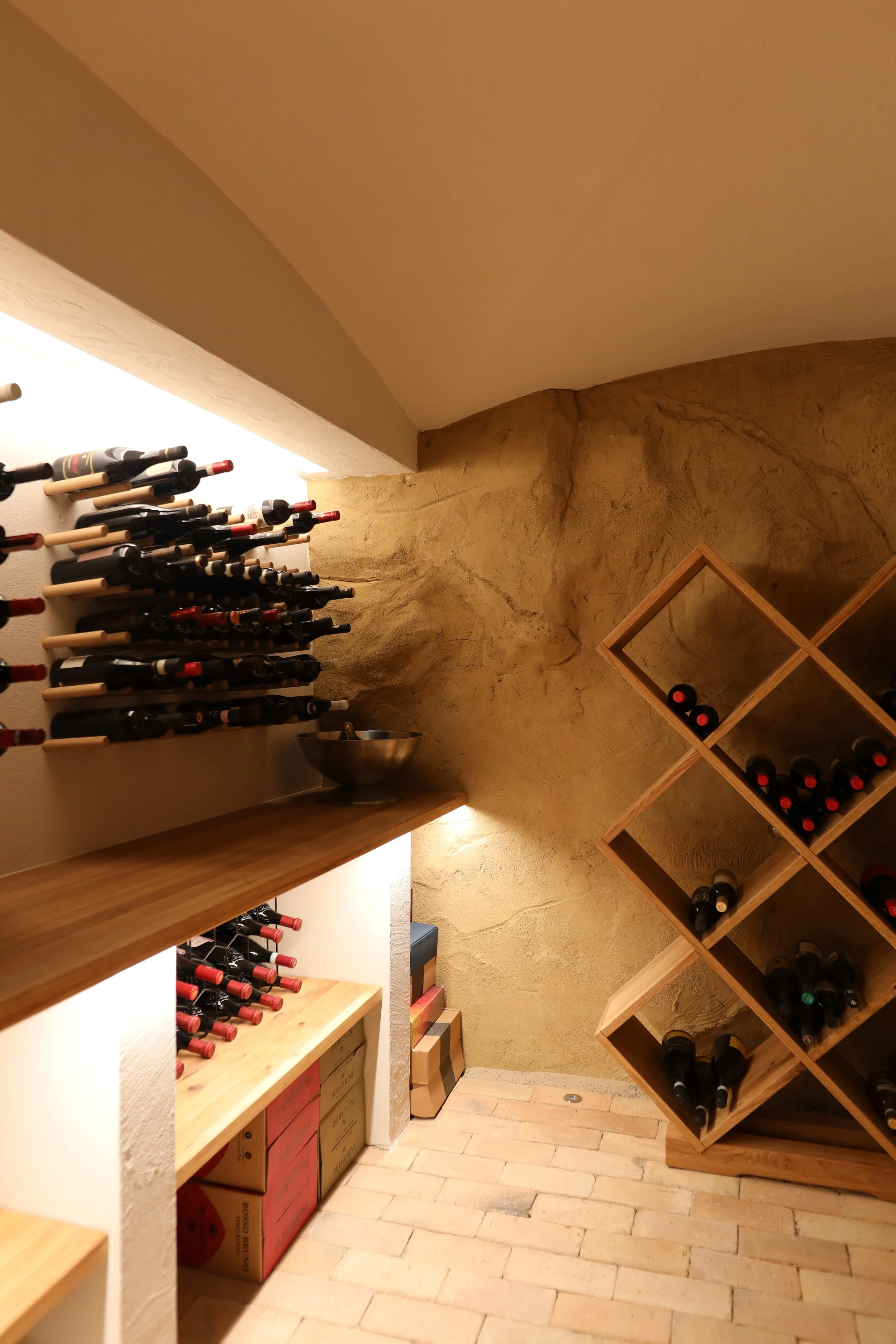 Ein Weinkeller mit Weinständern an der Wand, Weinflaschen auf Holzregalen und Brennholzplatten an der Wand.