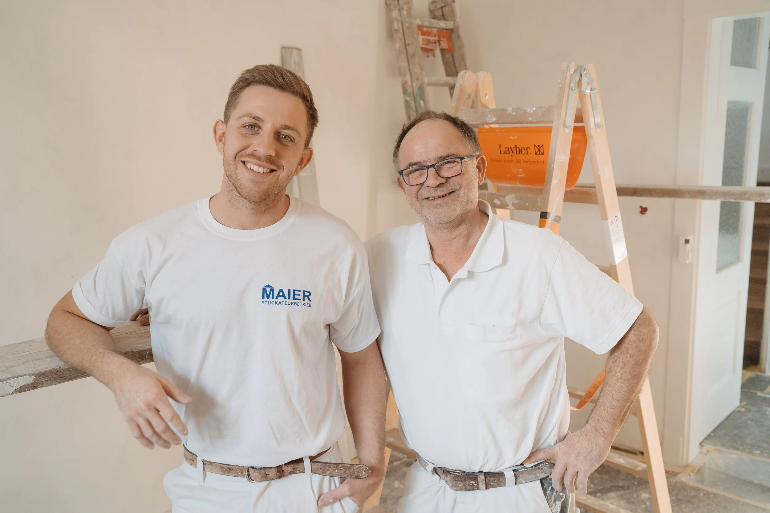 Zwei Männer stehen nebeneinander in einem Raum, der sich im Renovierungsprozess befindet, mit einer Leiter im Hintergrund. Beide tragen weiße T-Shirts, eines mit dem Logo 'Maier'.