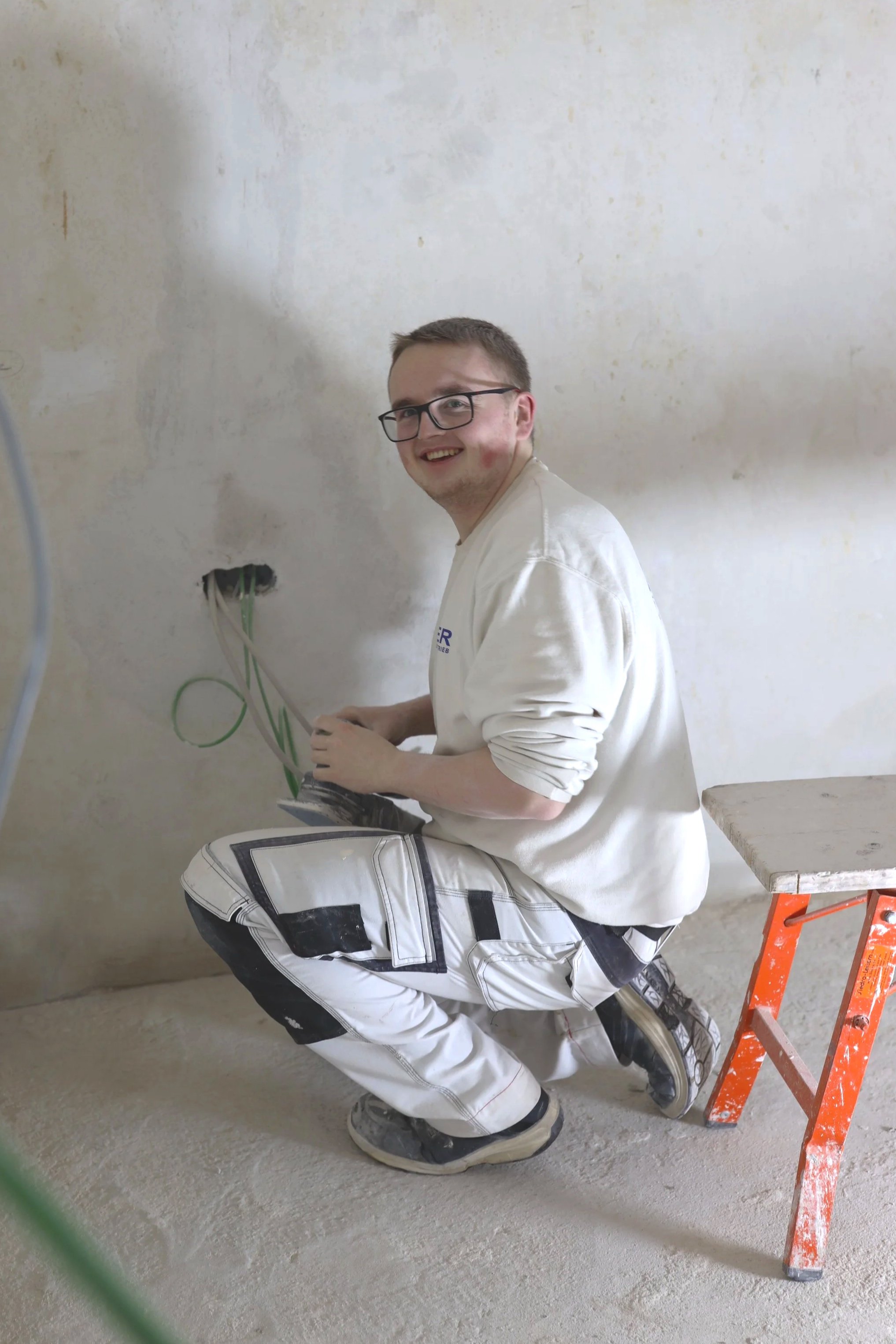 Ein junger Mann trägt eine weiße Arbeitsjacke und schwarze-weiße Hosen, arbeitet an einer Wand, sitzt auf dem Boden, trägt eine Brille und lächelt. Er ist neben einer orangefarbenen Leiter, die auf einer Baustelle steht.