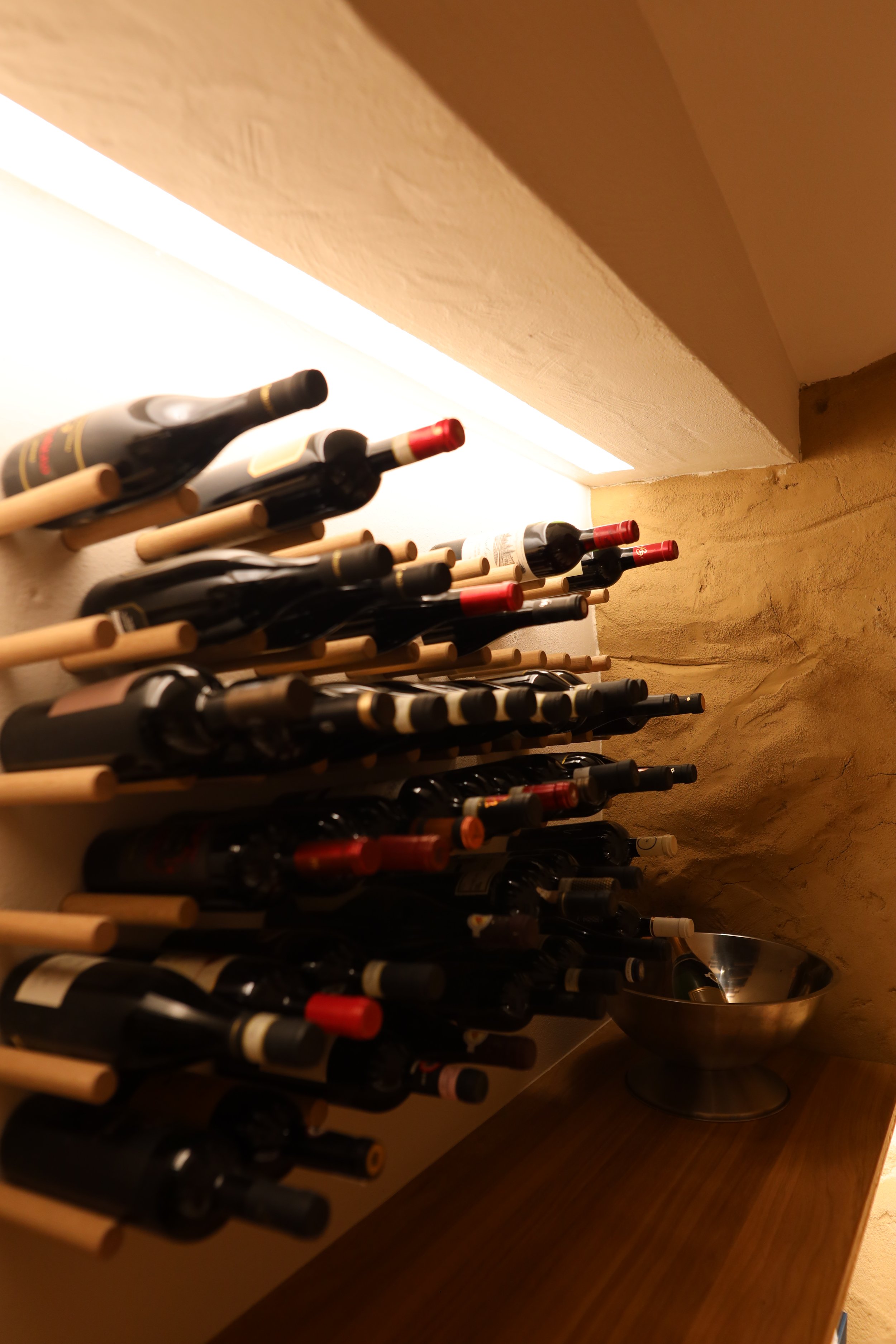 Blick auf eine Weinsammlung in einem Regal, mit mehreren Weinflaschen in Holzhaltern, vor einer Ziegelwand und einem Tisch mit einem silbernen Weinkühler.