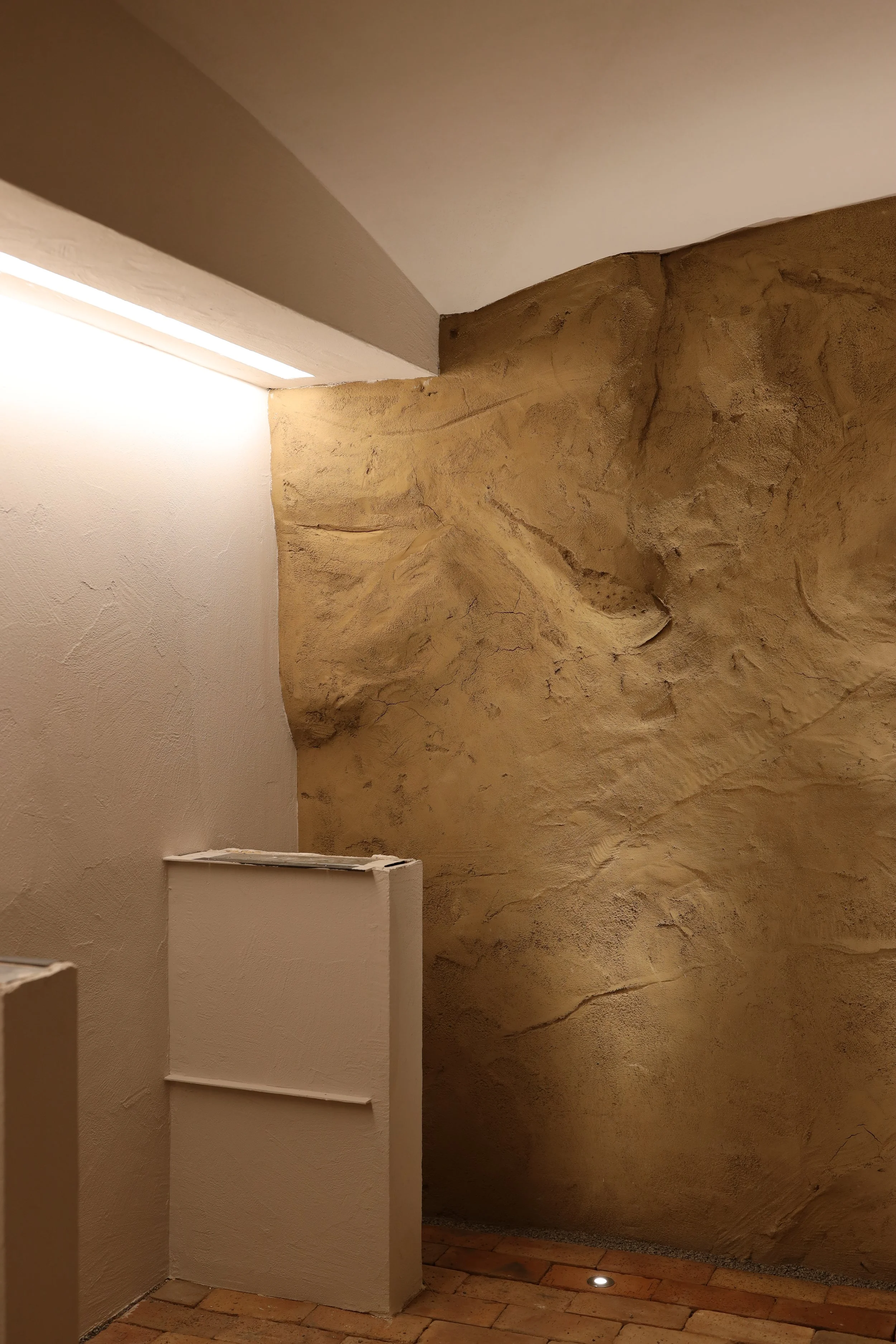 Blick auf eine Wand mit einer dekorativen Grobmauer-Struktur, beleuchtet durch indirektes Licht, mit weißen Zapfenregalen und terrakotta-farbigen Fliesen.