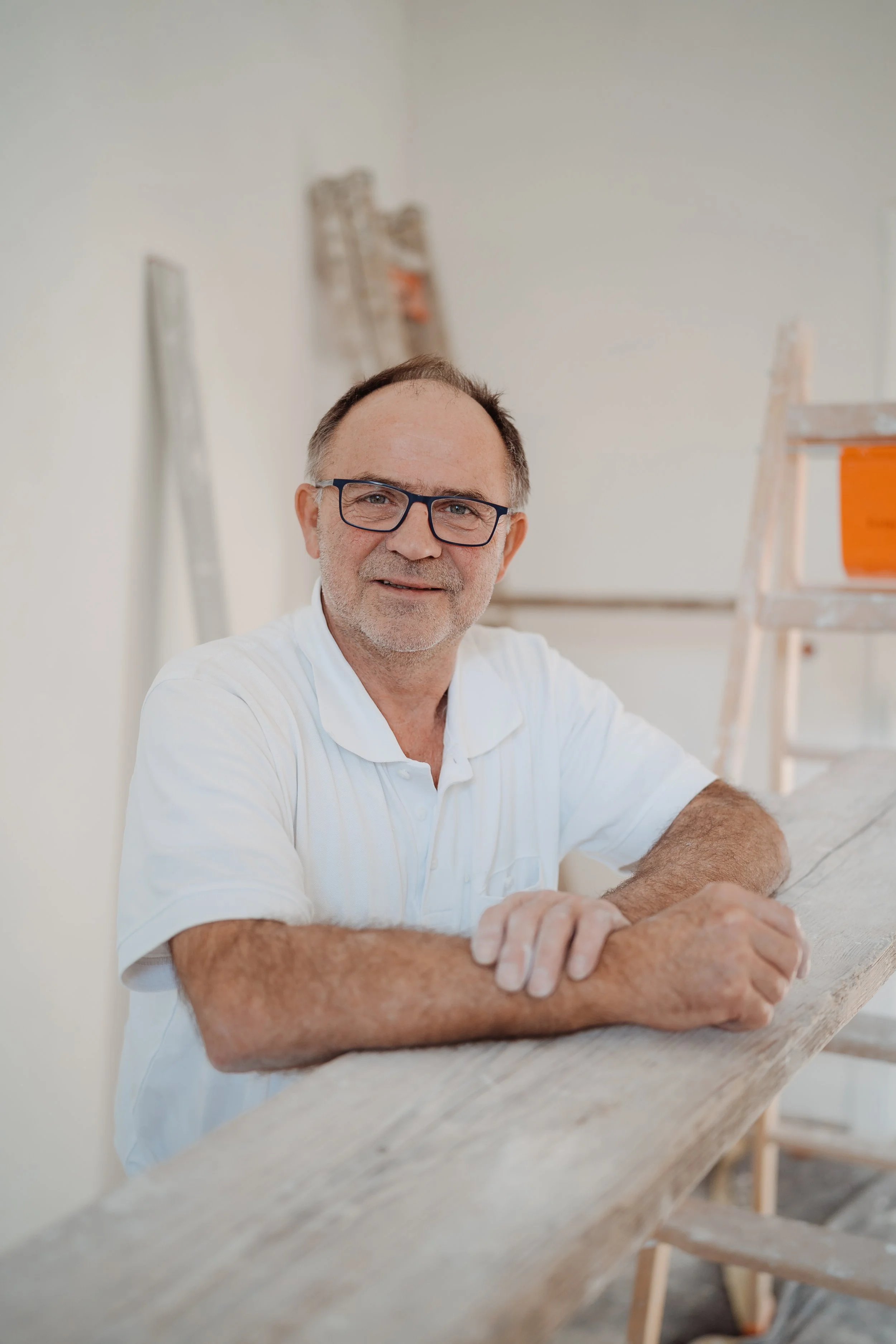 Ein älterer Mann mit Brille und Bart, der an einer Holzwerkbank an einem Projekt arbeitet, in einem Raum, der wahrscheinlich eine Werkstatt ist.