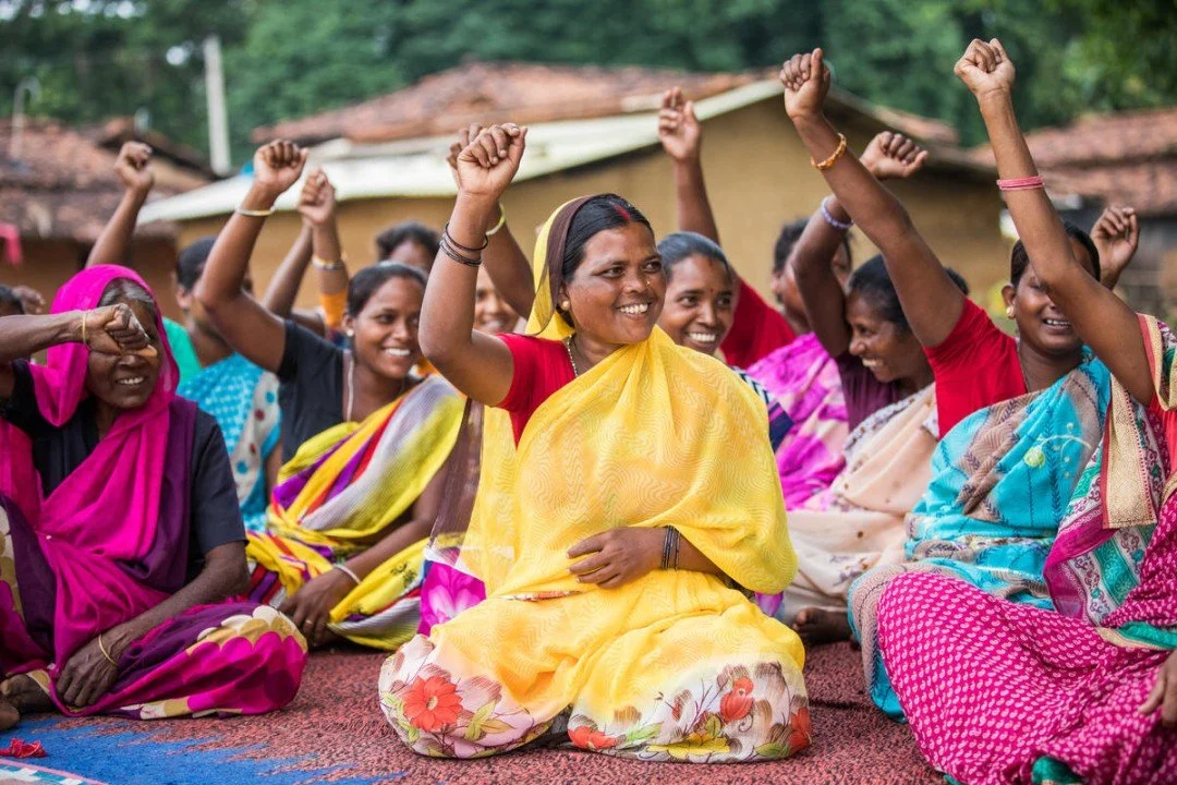 A group of women dressed in colorful traditional Indian sarees sitting on the ground outdoors, raising their hands in celebration and smiling.