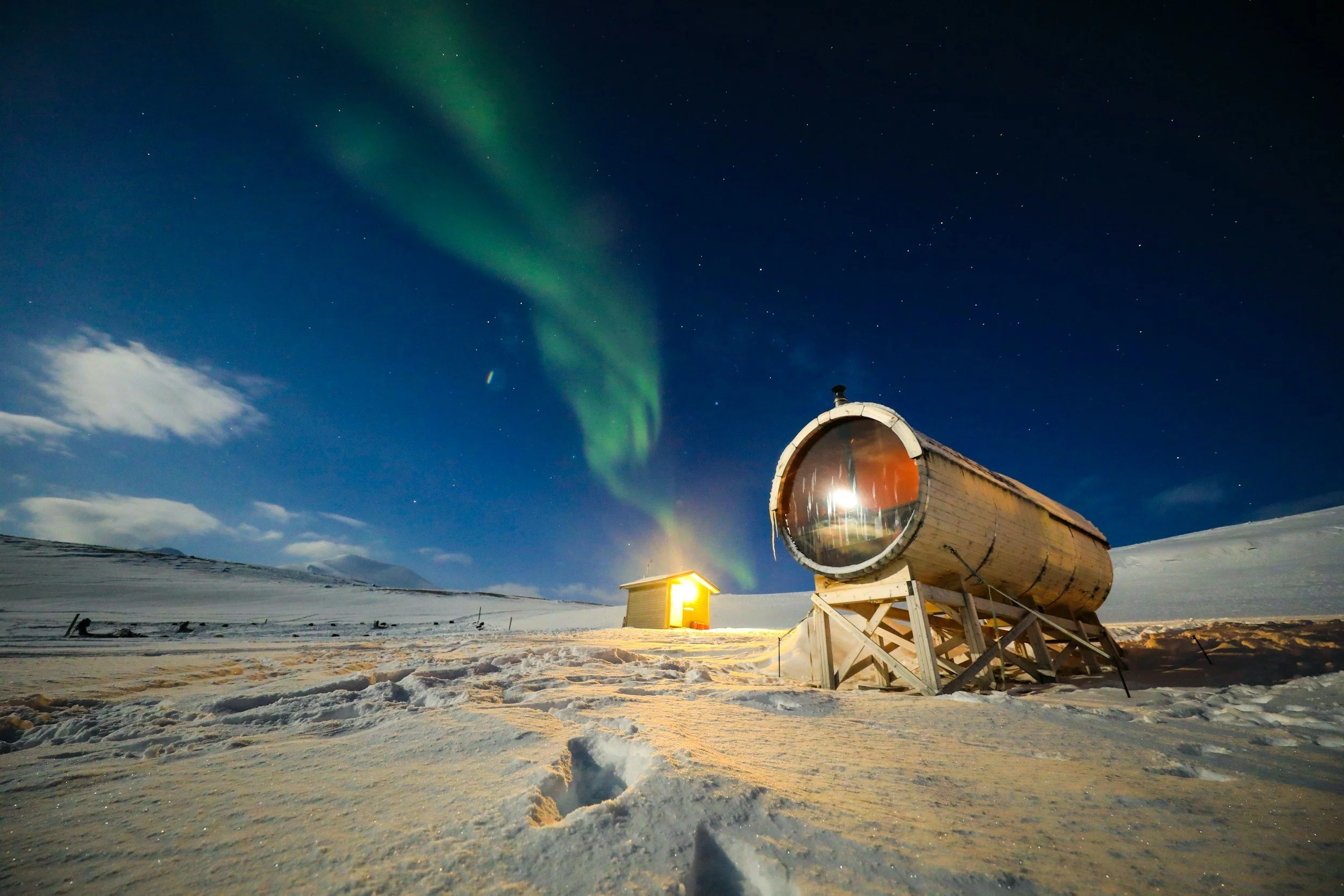 Northern lights in the night sky over a snow-covered landscape with a large telescope and a small illuminated building.