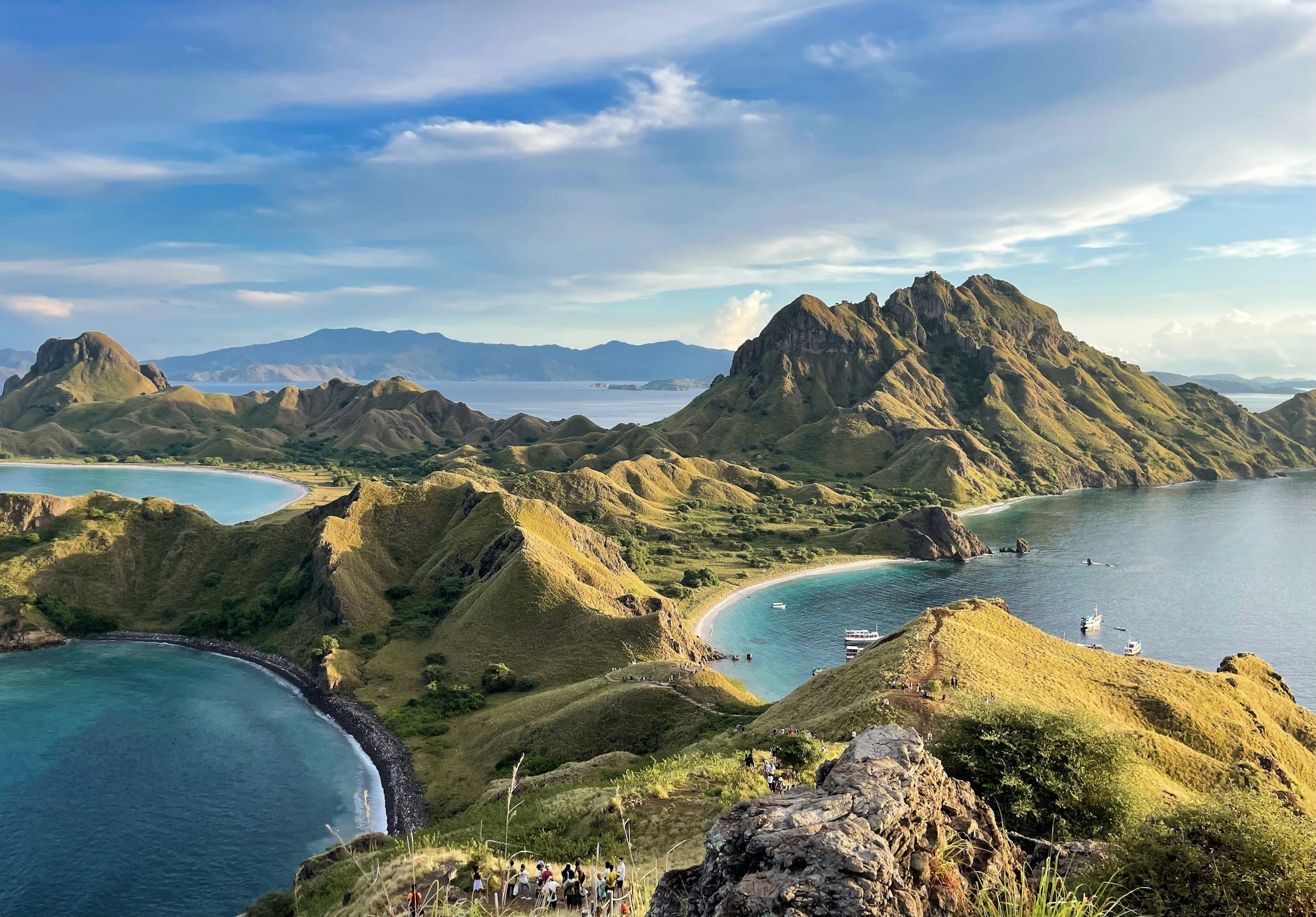 A scenic landscape with green, hilly terrain surrounding blue water, with mountains in the background and a partly cloudy sky.