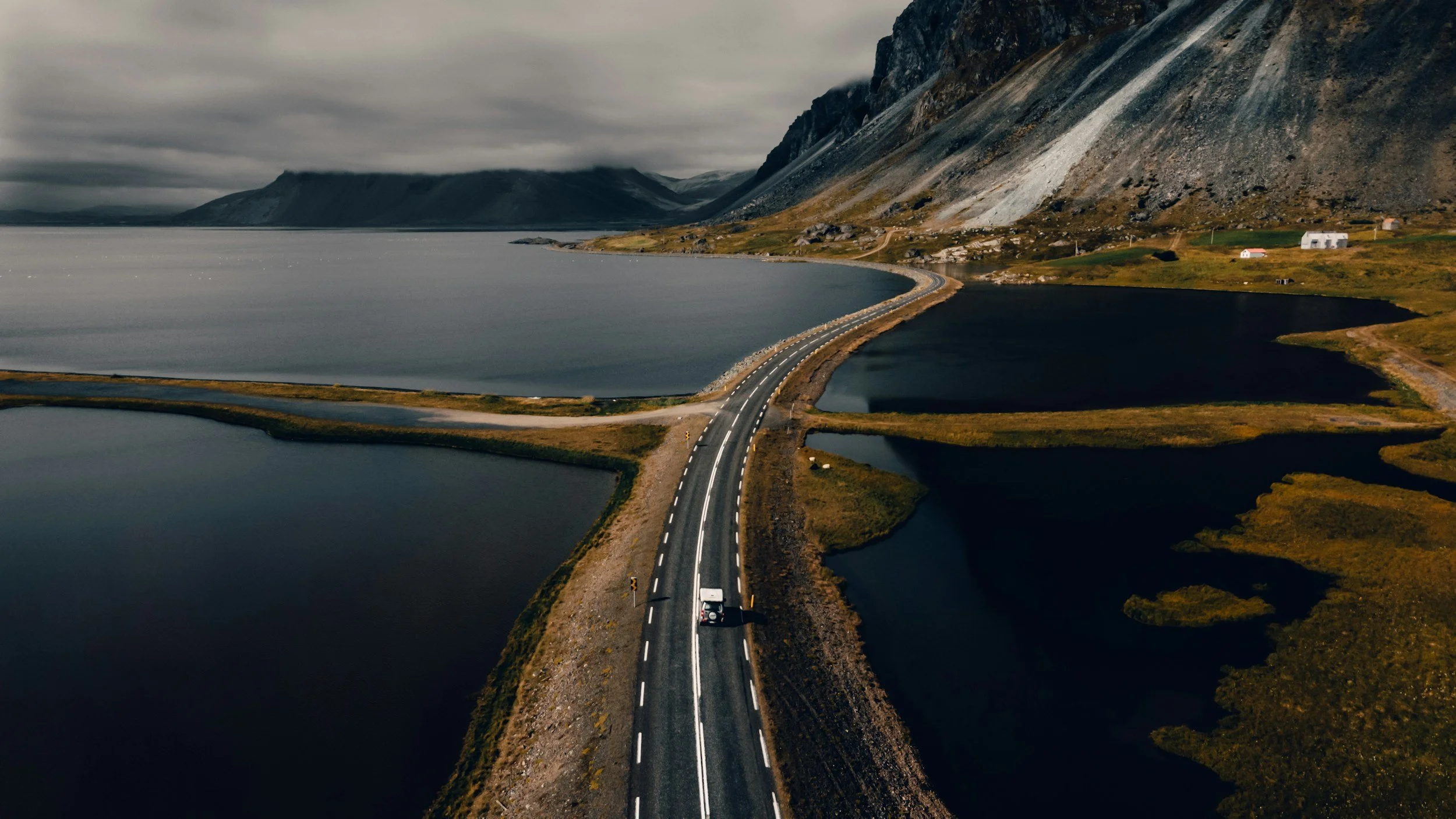 Aerial view of a narrow road crossing over a body of water, with mountains and cloudy sky in the background.