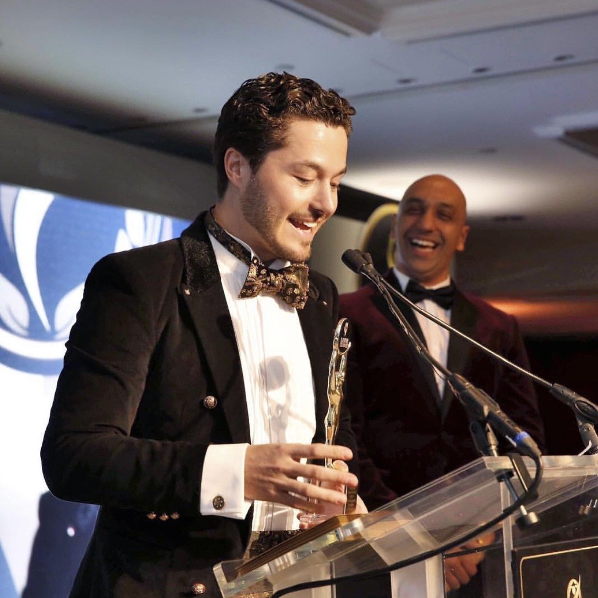 Two men in tuxedos, one holding an award and the other laughing, at an event with a microphone and a podium.