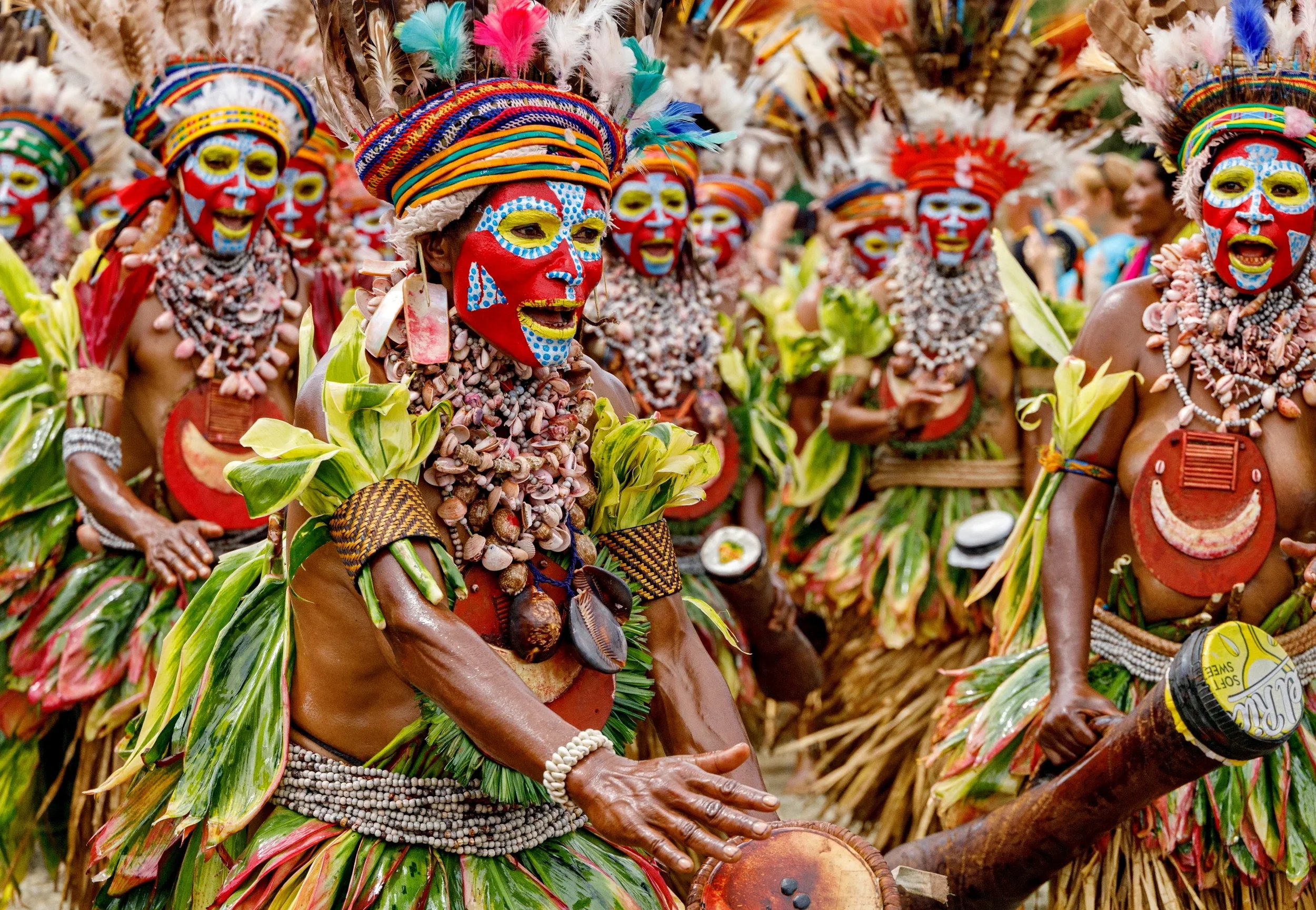 Group of people dressed in traditional colorful tribal costumes with face paint, feathers, shells, and leaf accessories, participating in a cultural festival or celebration.