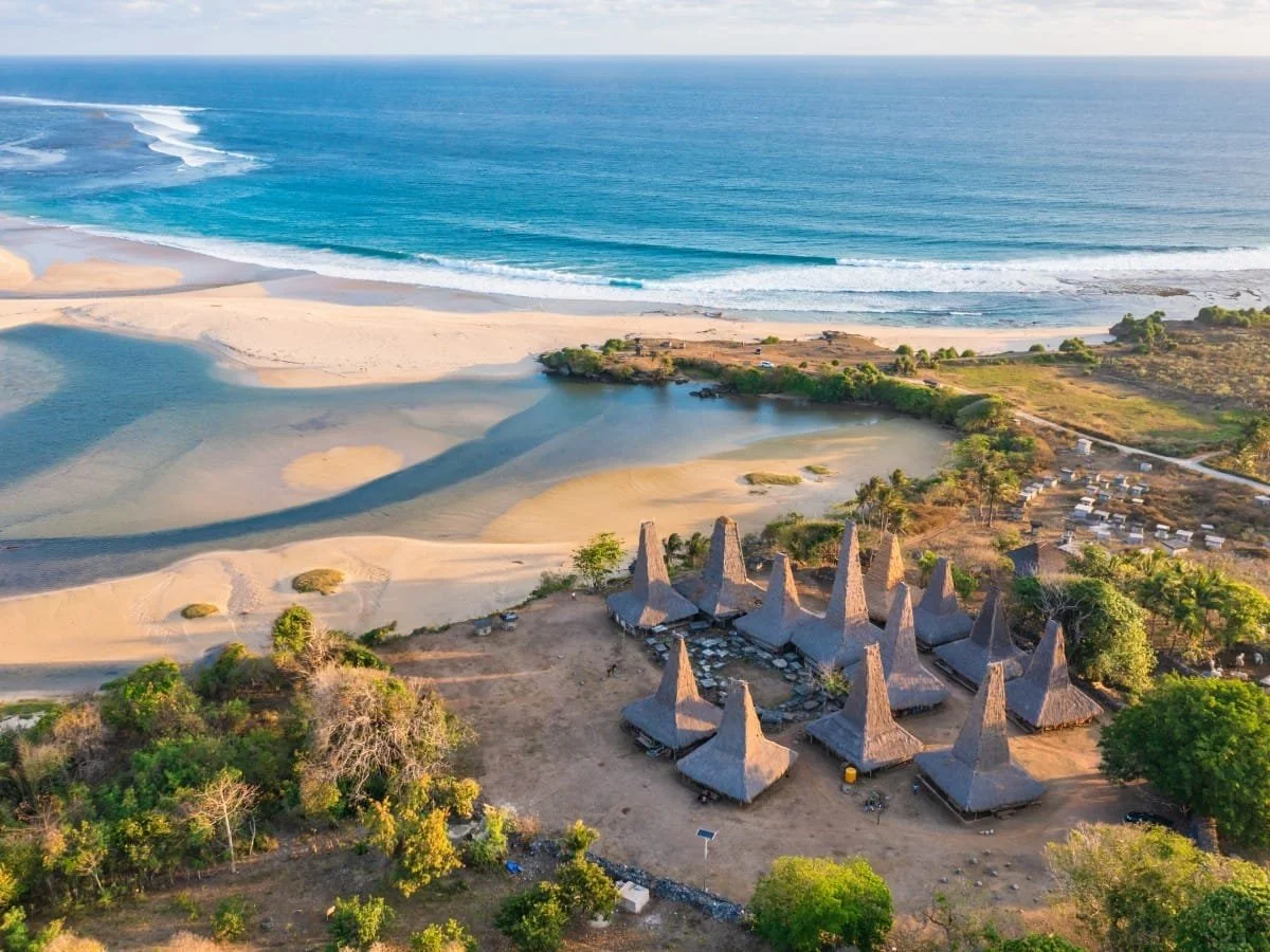 Aerial view of a beach with sand dunes, the ocean, and traditional thatched-roof structures near the shoreline, surrounded by green vegetation.