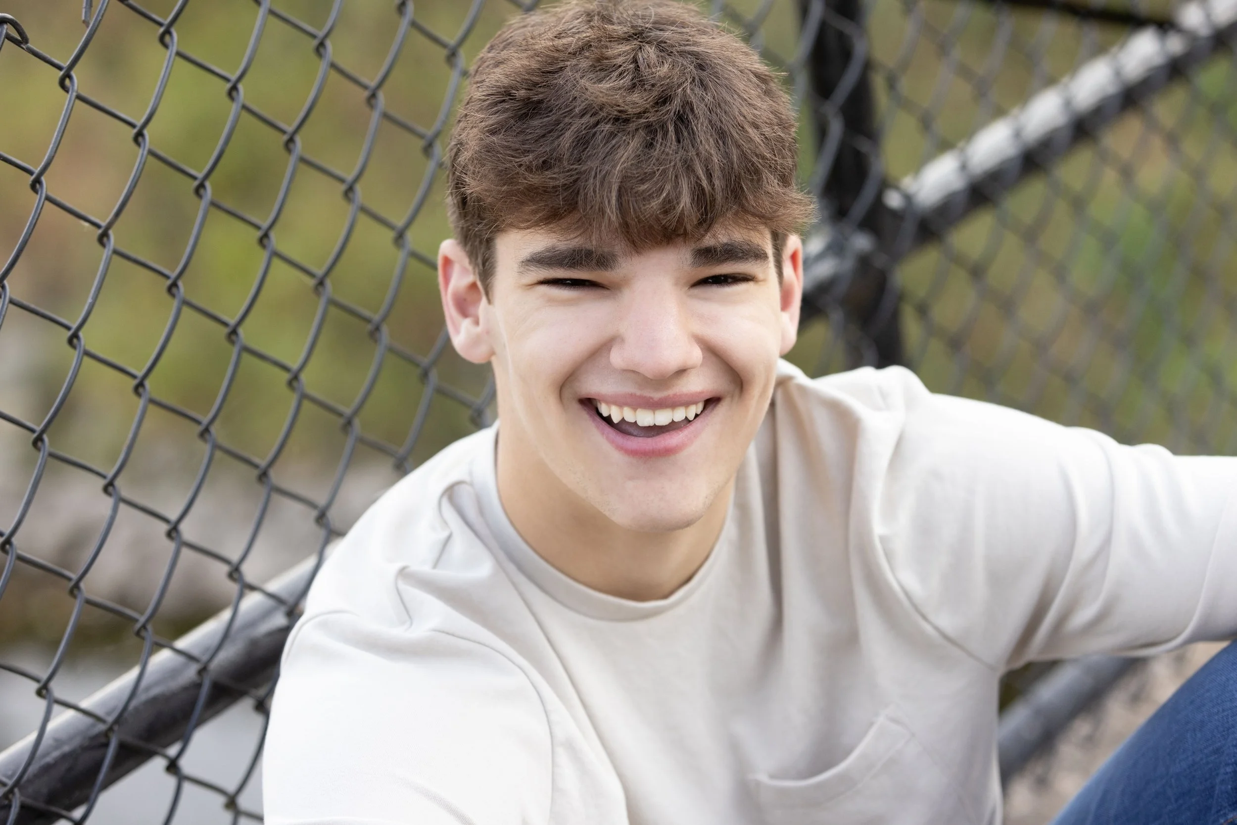 A young man with brown hair smiling and sitting outdoors near a chain-link fence.