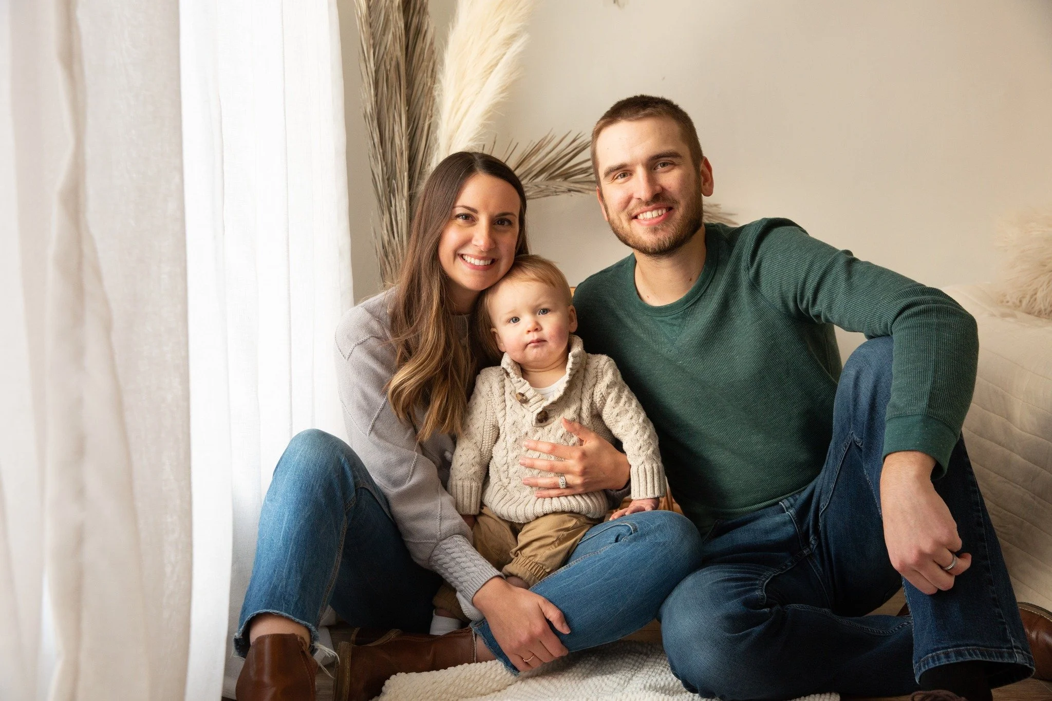 A smiling family of three sitting on the floor in a cozy, well-lit room. The woman has long brown hair, the man has short brown hair, and there is a young child with blond hair in the middle. They are all wearing casual clothing and appear happy.
