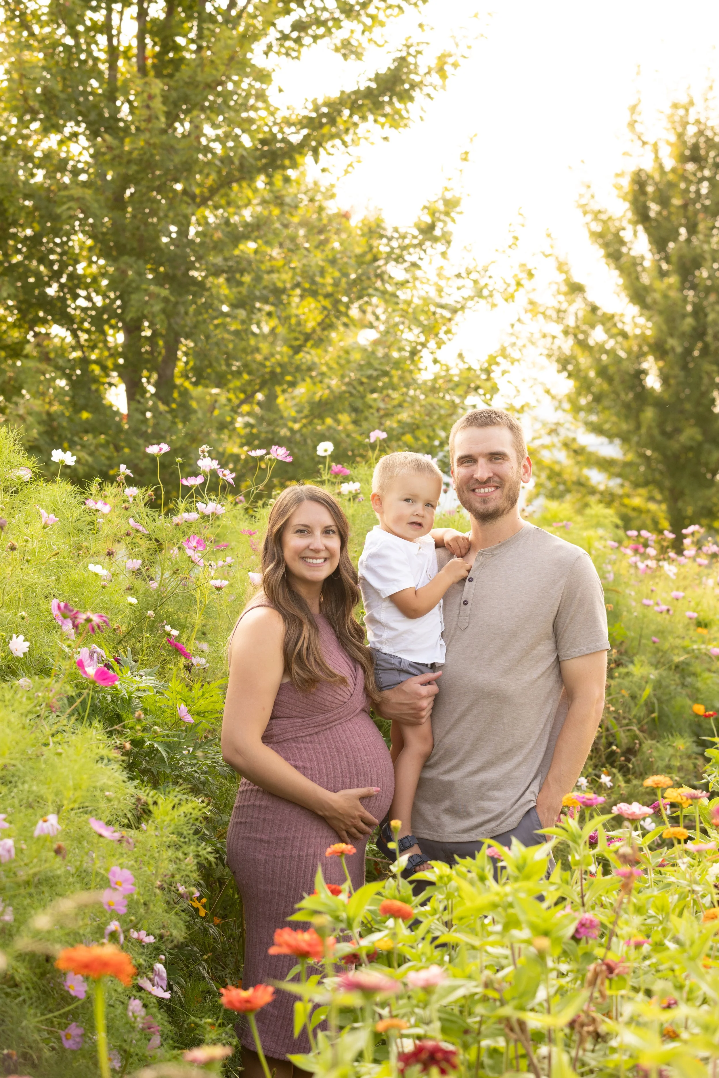 A family of three standing in a field of colorful flowers with green trees in the background during sunset, smiling at the camera. The woman is pregnant, the man is holding a young boy, all dressed casually.