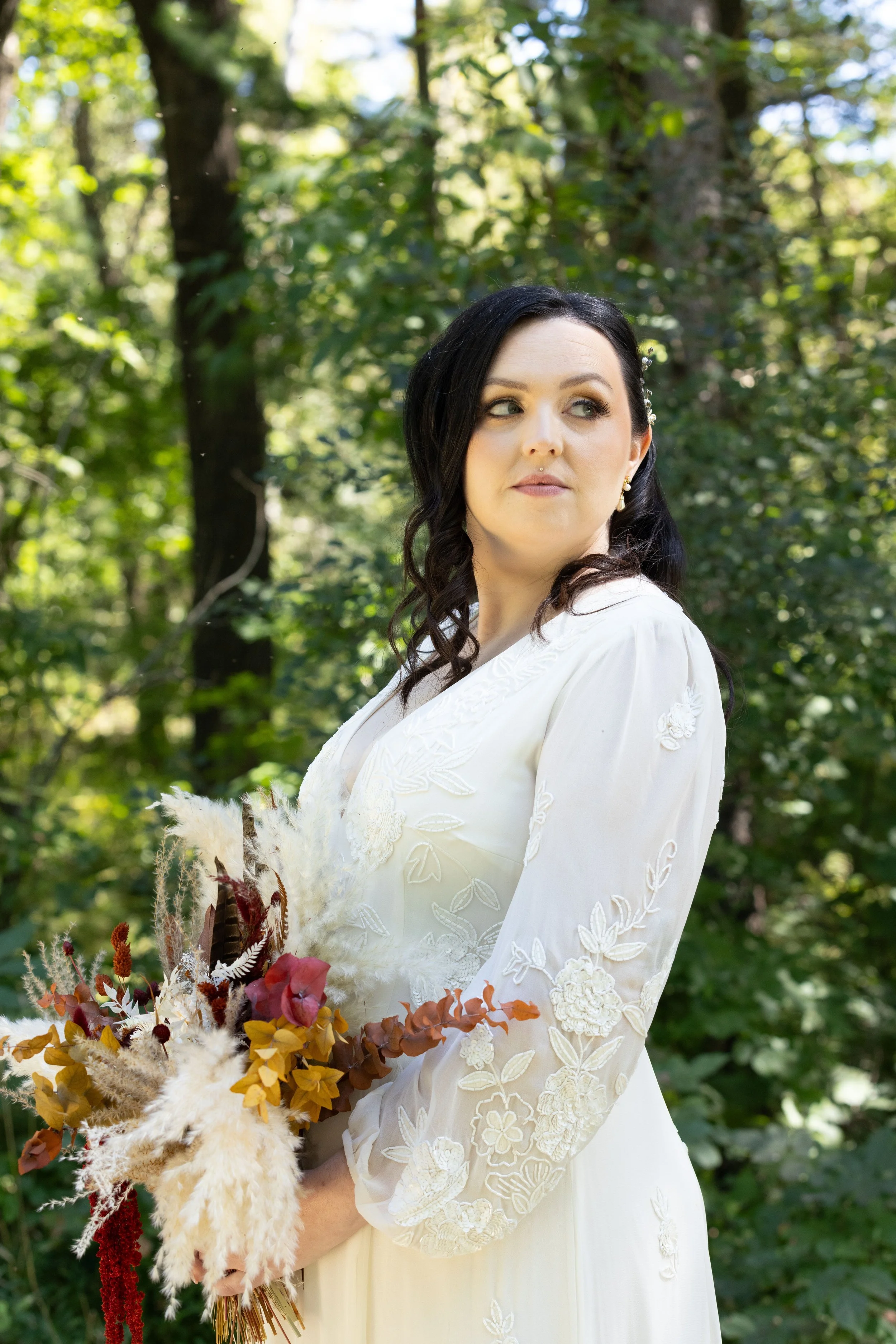 Woman in a white embroidered dress holding a bouquet of dried flowers in a green forest.