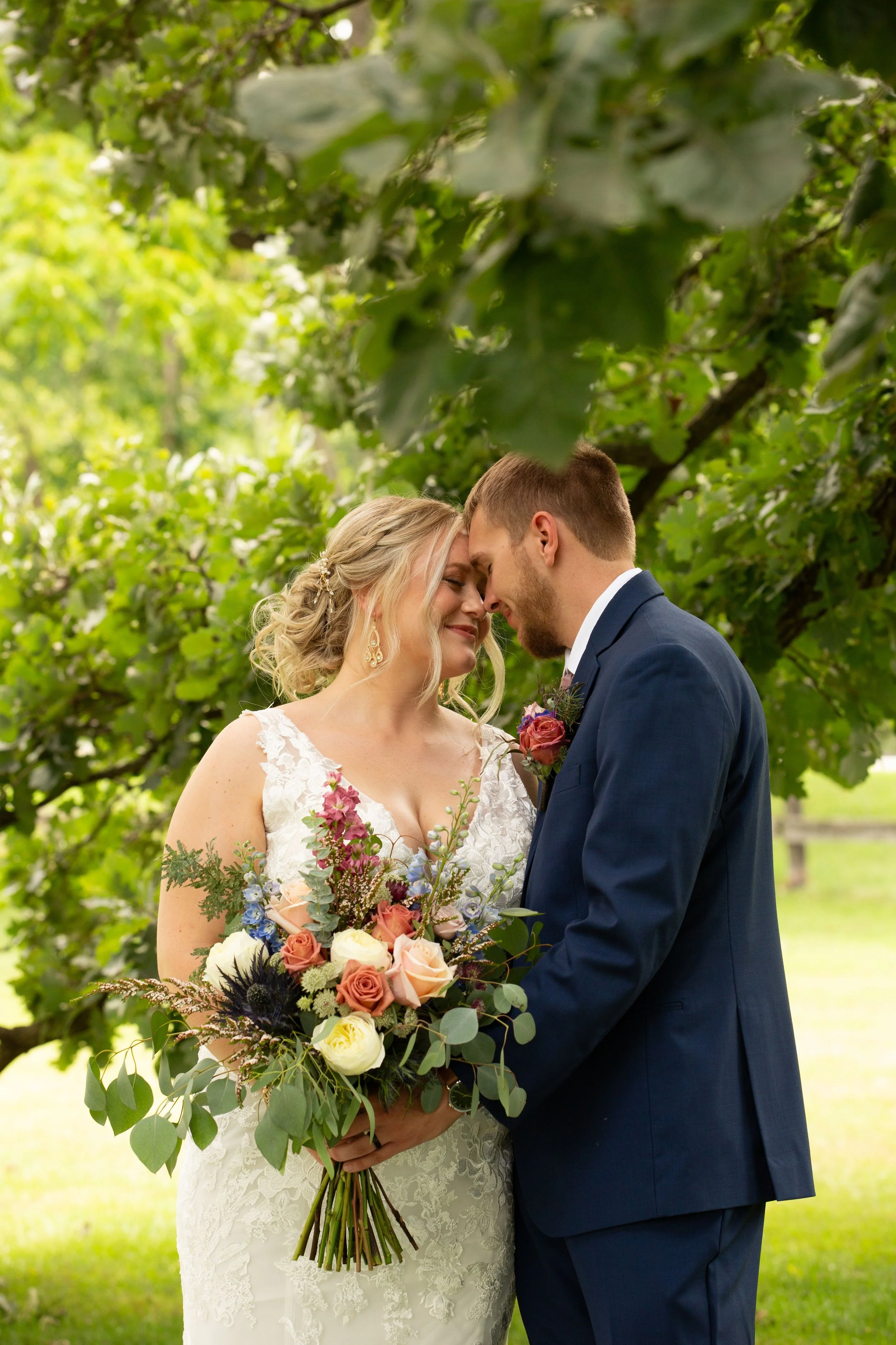 A bride and groom embrace outdoors under green trees, with the bride holding a bouquet of flowers.